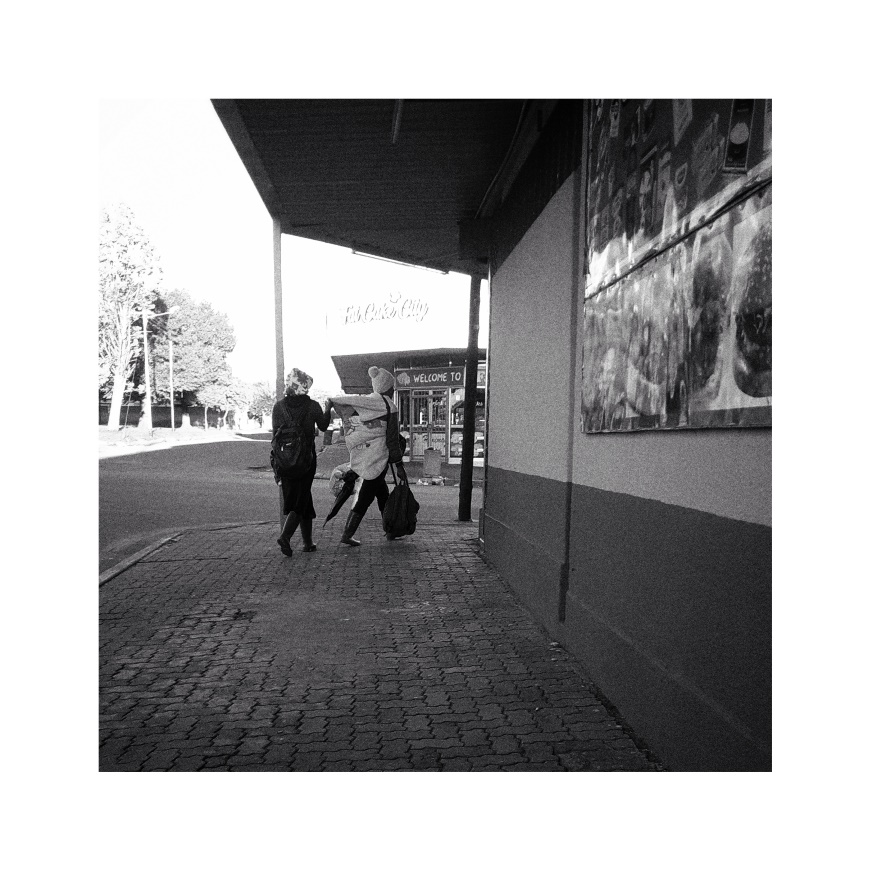Two women walking on a sidewalk. One of the women is carrying a baby on her back wrapped in a blanket.