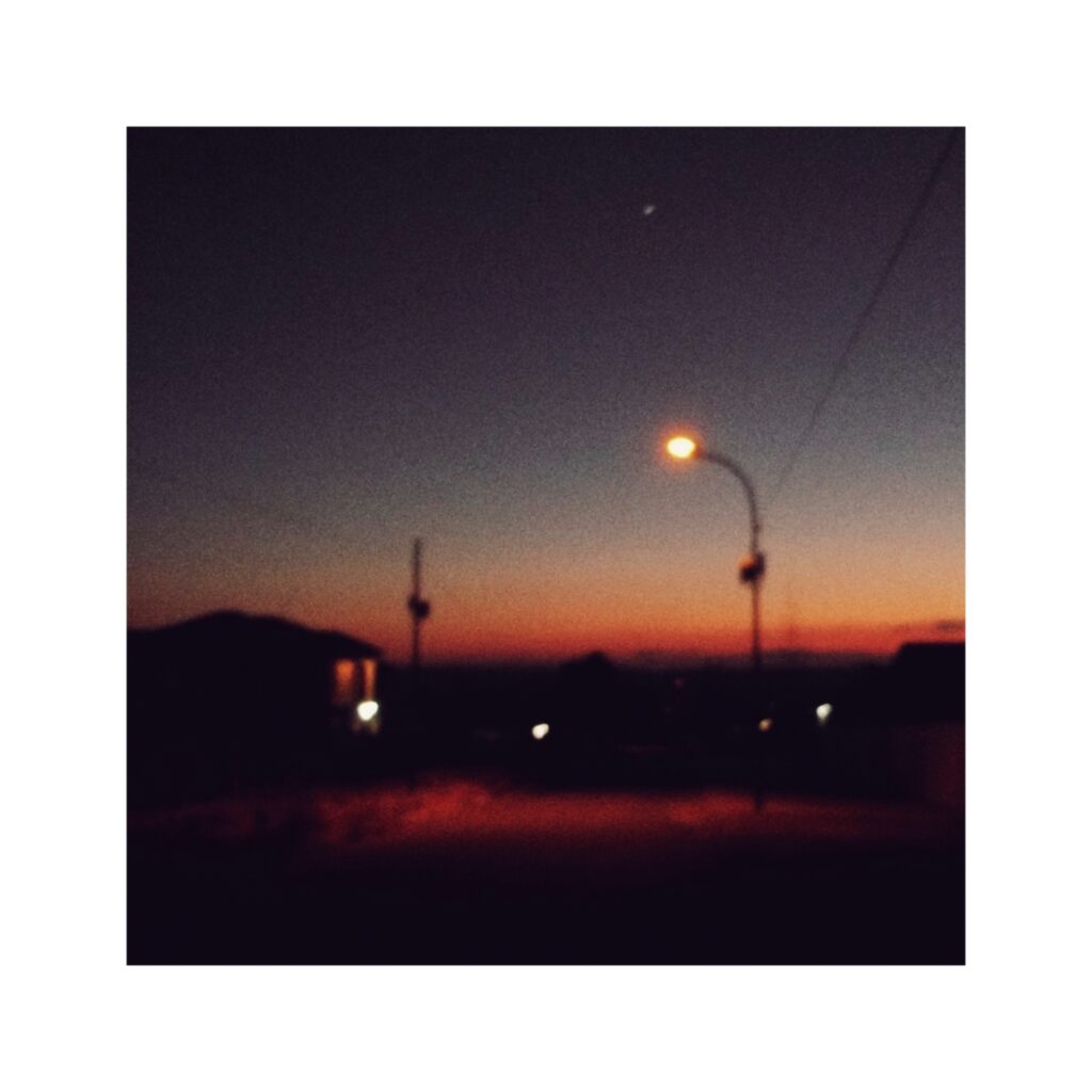 A photo of a street lamp in the foreground. In the distance a hazy horizon just before sunrise. The dark sky on top and the pink red and orange of the sun in the bottom third of the image.